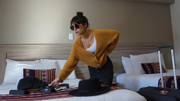 A woman wearing round sunglasses and a mustard yellow cardigan leaning over a hotel bed to organize travel accessories, including a black hat and camera, next to a suitcase.