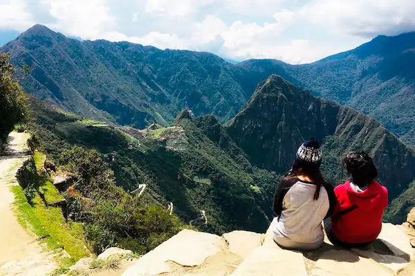 Two hikers sitting on a stone ledge overlooking the ancient ruins of Machu Picchu and surrounding green Andean mountains along the Inca Trail